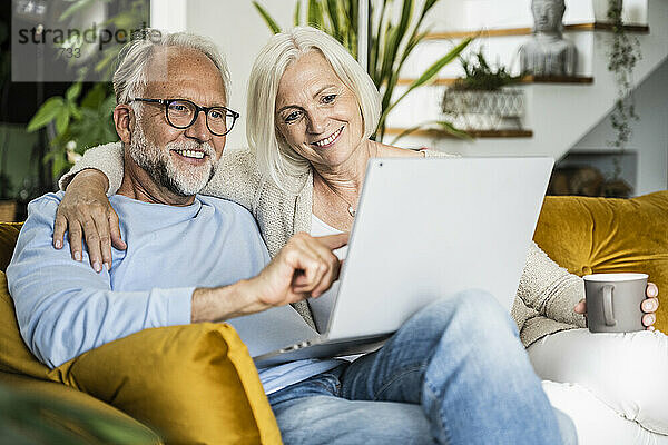 Lächelnder reifer Mann  der einen Laptop benutzt  während er mit einer Frau auf dem Sofa zu Hause sitzt