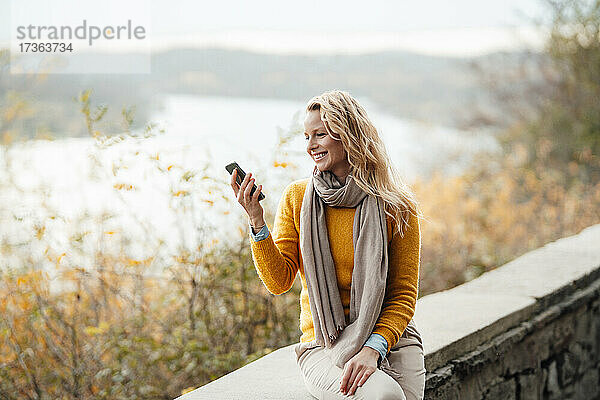 Lächelnde blonde Frau  die während eines Videogesprächs über ein Mobiltelefon auf einer Stützmauer sitzt