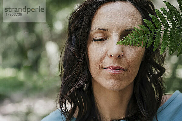 Frau mit geschlossenen Augen hält Farnblatt im Wald