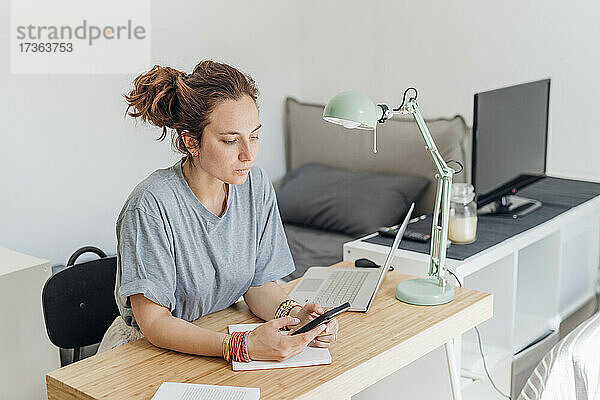 Freiberuflerin  die ein Mobiltelefon benutzt  während sie mit einem Laptop in ihrer Wohnung sitzt