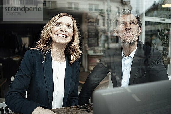 Lächelnde blonde ältere Geschäftsfrau  die mit einem männlichen Kollegen in einem Café sitzt  gesehen durch ein Glasfenster