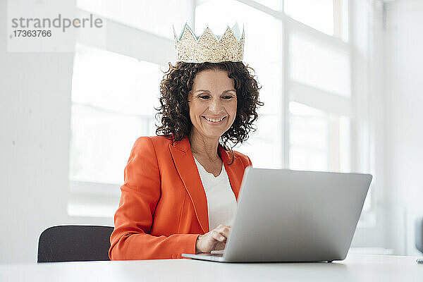 Lächelnde weibliche Fachkraft  die eine Krone trägt  während sie einen Laptop im Büro benutzt