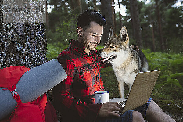 Lächelnder männlicher Wanderer  der einen Laptop benutzt  während er mit seinem Hund im Wald sitzt