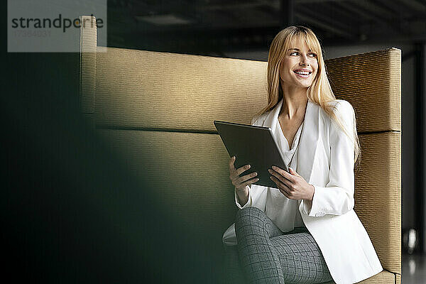 Lächelnde Geschäftsfrau schaut weg  während sie mit einem digitalen Tablet auf dem Sitz sitzt