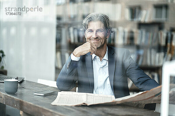 Lächelnder Geschäftsmann mit Zeitung im Café sitzend durch Glasfenster gesehen