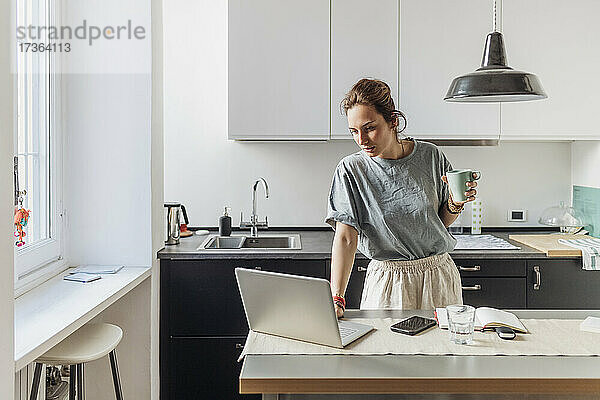 Frau hält Kaffeetasse und benutzt Laptop in der Küche