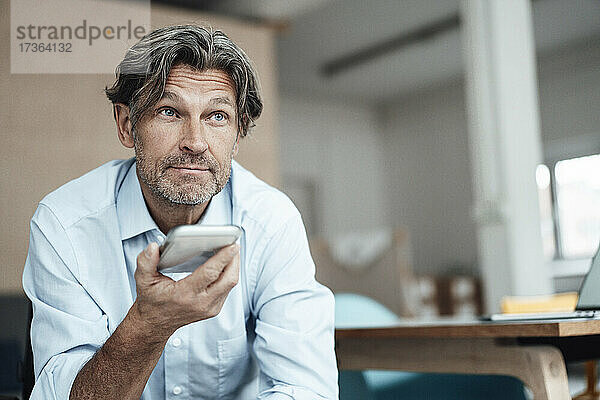 Älterer Geschäftsmann  der im Büro sitzend mit seinem Smartphone telefoniert