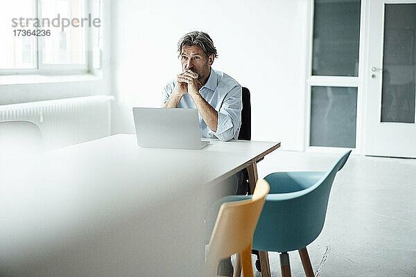 Älterer Geschäftsmann sitzt mit verschränkten Händen am Schreibtisch im Büro