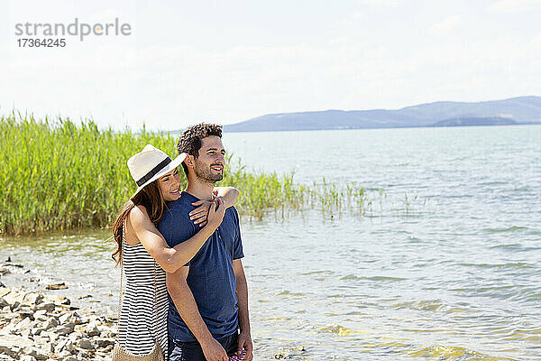 Lächelnde Frau mit Hut  die einen Mann von hinten umarmt  während sie am Trasimeno-See steht