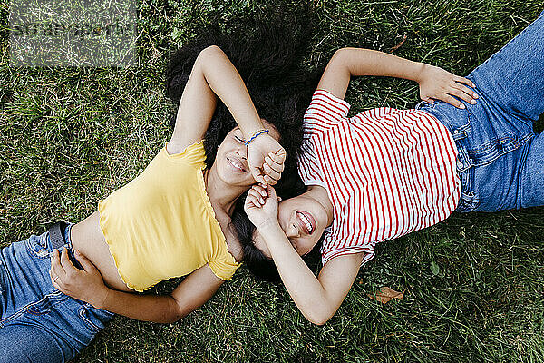 Lächelndes lesbisches Paar  das die Augen mit den Händen bedeckt  während es im Park im Gras liegt