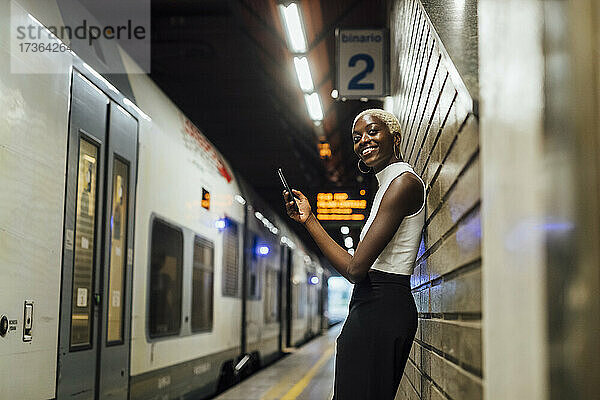 Lächelnde Frau mit Mobiltelefon an der Wand lehnend in einer U-Bahn-Station