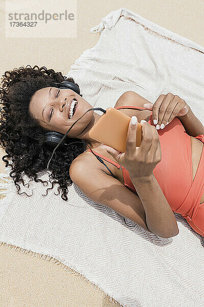 Fröhliche Frau mit geschlossenen Augen hört Musik  während sie auf einem Strandtuch liegt