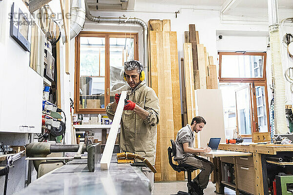 Älterer männlicher Tischler bei der Arbeit an Holz  während ein Kollege in der Werkstatt schreibt