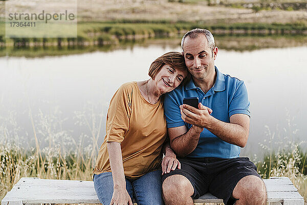 Sohn benutzt sein Smartphone  während er mit seiner Mutter auf einer Bank am See sitzt