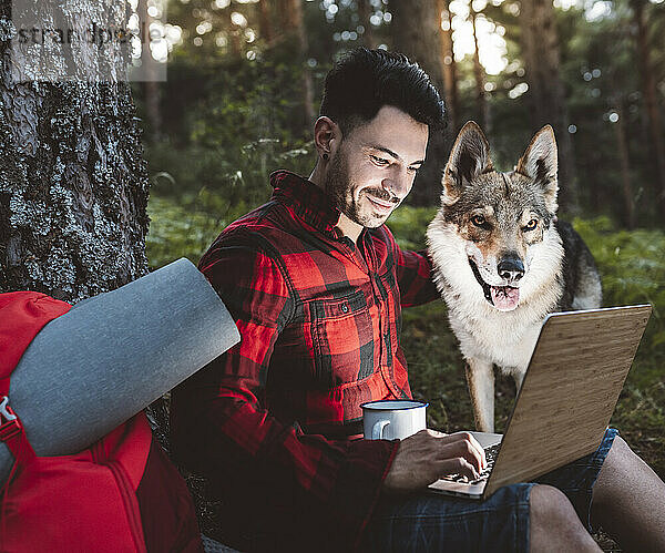 Mittlerer erwachsener Mann arbeitet an seinem Laptop  während er mit seinem Hund im Wald sitzt