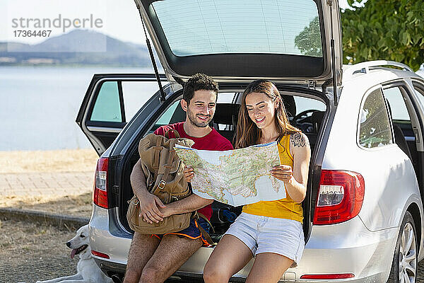 Lächelndes Paar  das im Kofferraum des Autos sitzend die Karte überprüft