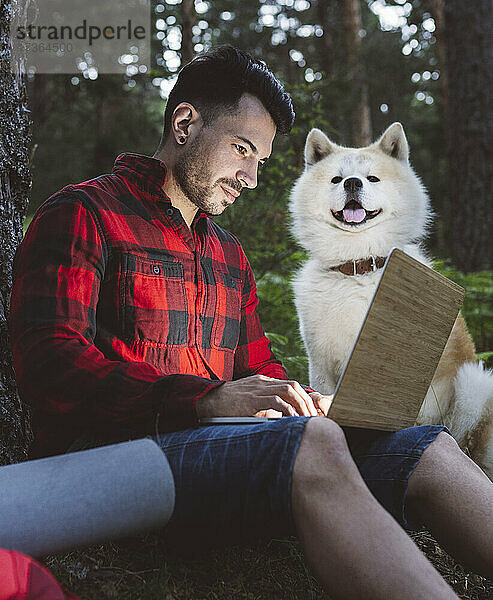 Mann arbeitet am Laptop  während er neben seinem Haustier im Wald sitzt