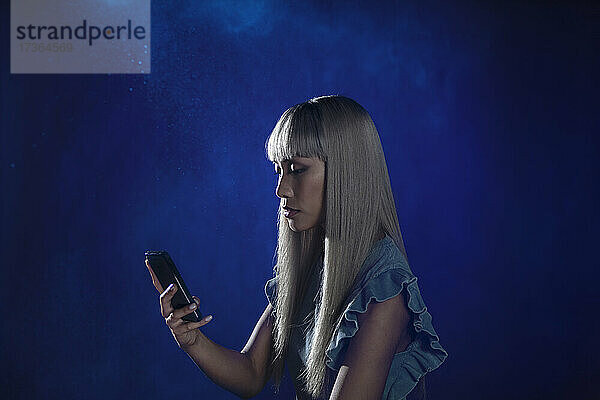 Junge Frau mit Pony benutzt ein Smartphone vor blauem Hintergrund