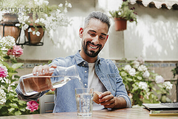 Lächelnder Geschäftsmann gießt am Schreibtisch Wasser in ein Glas