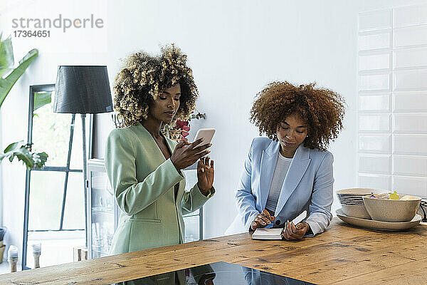 Junge Geschäftsfrau  die ein Smartphone benutzt  während eine Kollegin den Terminkalender auf dem Tisch betrachtet