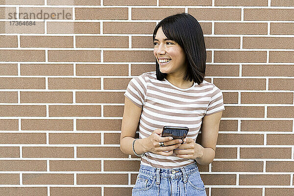 Lächelnde junge Frau mit Handy in der Hand  die sich an eine Backsteinmauer lehnt