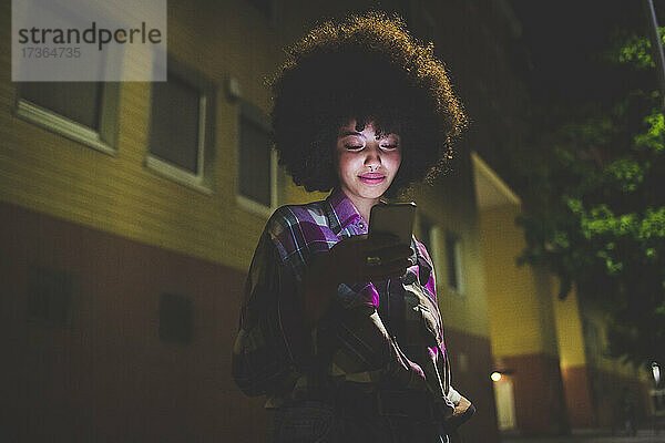 Junge Frau mit Afrofrisur  die nachts in der Stadt ihr Smartphone benutzt