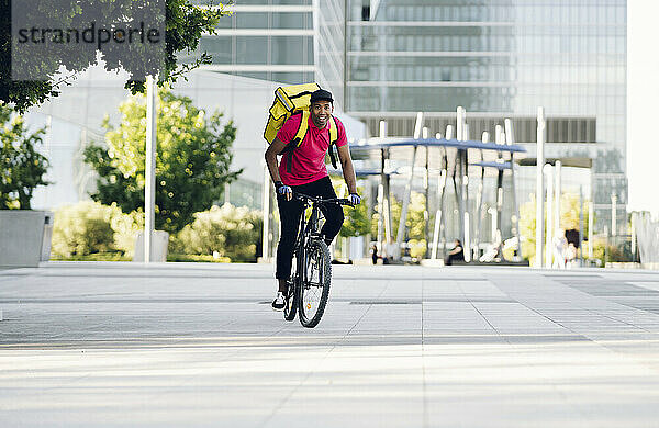 Lächelnder Zusteller mit gelbem Rucksack auf dem Fahrrad in der Stadt