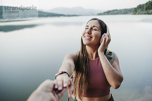 Lächelnde junge Frau mit geschlossenen Augen hält die Hand eines Freundes am Seeufer