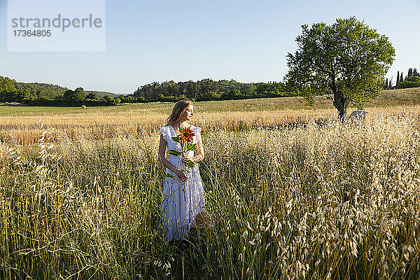 Junge Frau hält Sonnenblume  während sie inmitten von Gras im Feld steht