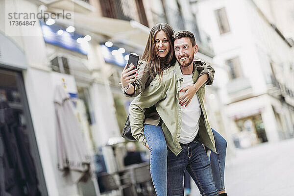 Lächelnde Frau  die ein Selfie mit ihrem Smartphone macht  während sie ihren Freund in der Stadt huckepack nimmt