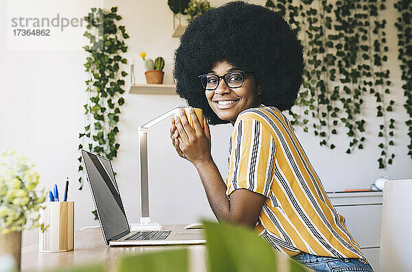 Lächelnde Afro-Frau  die eine Kaffeetasse hält  während sie am Schreibtisch sitzt