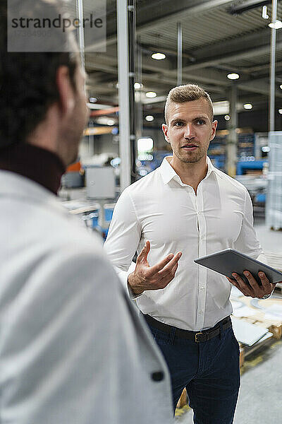Geschäftsmann hält ein digitales Tablet in der Hand  während er sich mit einem Kollegen in einer Fabrik unterhält
