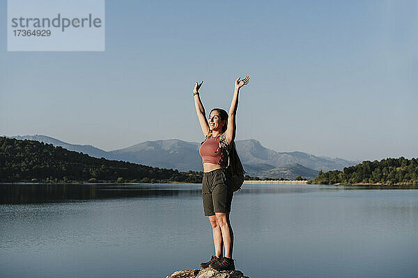 Lächelnde Wanderin mit erhobenen Armen auf einem Felsen am See stehend