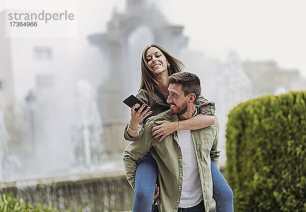 Lächelnde Frau  die ein Mobiltelefon benutzt  während sie auf ihrem Freund huckepack sitzt