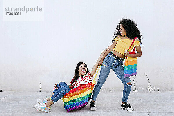 Junge lesbische Frau  die ihre Freundin in einer Tasche mit Regenbogenfahne auf dem Gehweg sitzend zieht