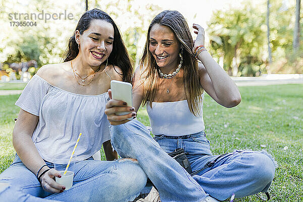 Lächelnde Frau  die mit ihrer Freundin im Park sitzt und ein Smartphone benutzt