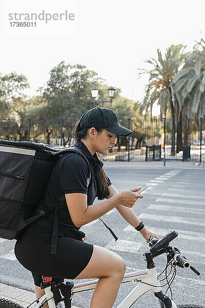 Junge Zustellerin  die auf dem Fahrrad sitzend ein Mobiltelefon benutzt