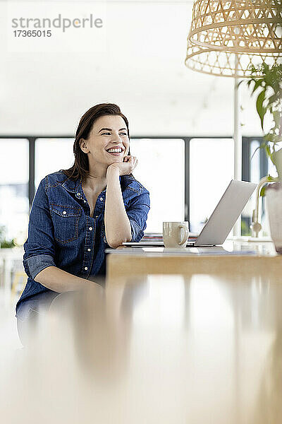 Nachdenkliche Geschäftsfrau mit Hand am Kinn am Laptop sitzend am Arbeitsplatz