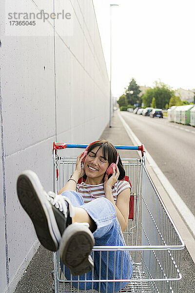 Lächelnde Frau mit geschlossenen Augen hört Musik im Einkaufswagen