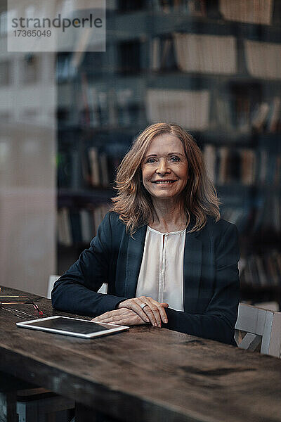 Lächelnde blonde Geschäftsfrau sitzt mit digitalem Tablet in einem Café  gesehen durch ein Glasfenster