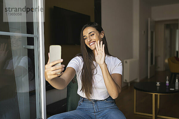 Lächelnde junge Frau winkt mit der Hand während eines Videoanrufs über ein Smartphone