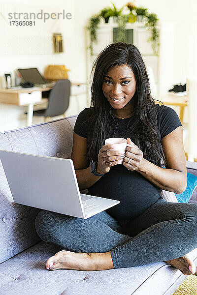 Lächelnde schwangere Frau sitzt mit Kaffeetasse und Laptop auf dem Sofa zu Hause