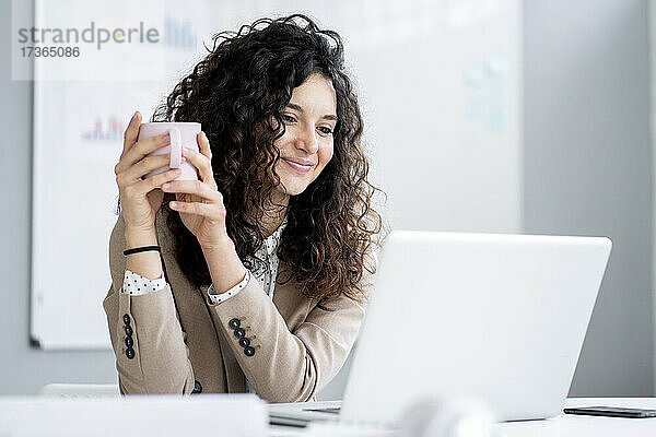 Geschäftsfrau mit Kaffeetasse  die lächelnd auf ihren Laptop schaut
