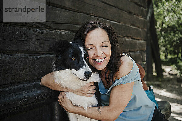 Lächelnde erwachsene Frau mit geschlossenen Augen  die einen Border Collie vor einem Haus umarmt