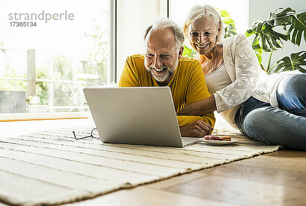 Lächelndes Paar  das einen Laptop benutzt  während es sich auf einem Teppich zu Hause entspannt