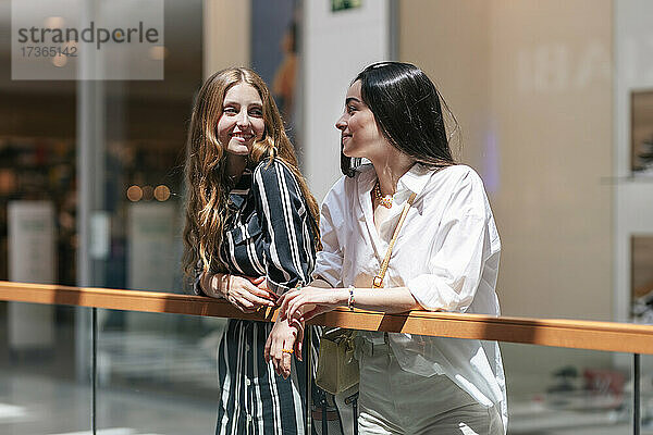 Lächelnde Freundinnen  die sich gegenseitig ansehen  während sie sich an ein Geländer im Einkaufszentrum lehnen