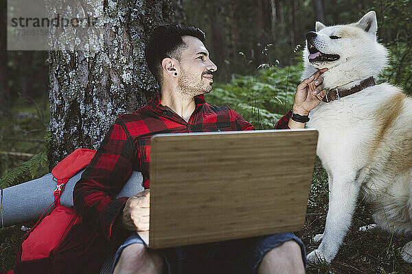 Lächelnder Mann mit Laptop  der einen Hund streichelt und ihn betrachtet  während er im Wald sitzt