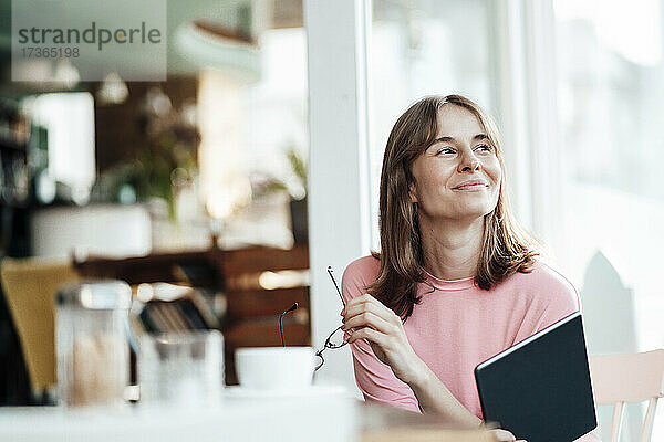 Lächelnde weibliche Fachkraft  die mit einem digitalen Tablet in einem Café sitzt und wegschaut