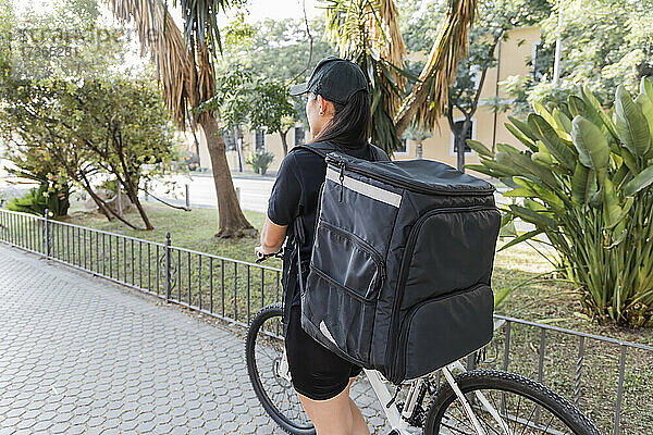 Weiblicher Zusteller  der einen Rucksack trägt  während er mit dem Fahrrad auf dem Gehweg fährt