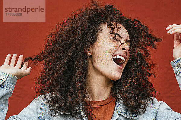 Fröhliche junge Frau mit lockigem Haar singt vor einer roten Wand
